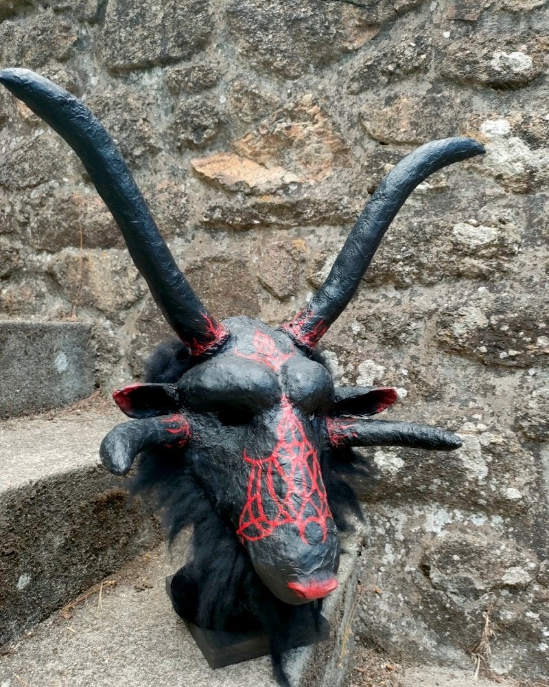 Máscara Asmodan, cabeza de macho cabrío negra de cuernos largos, con una telaraña roja pintada en el hocico